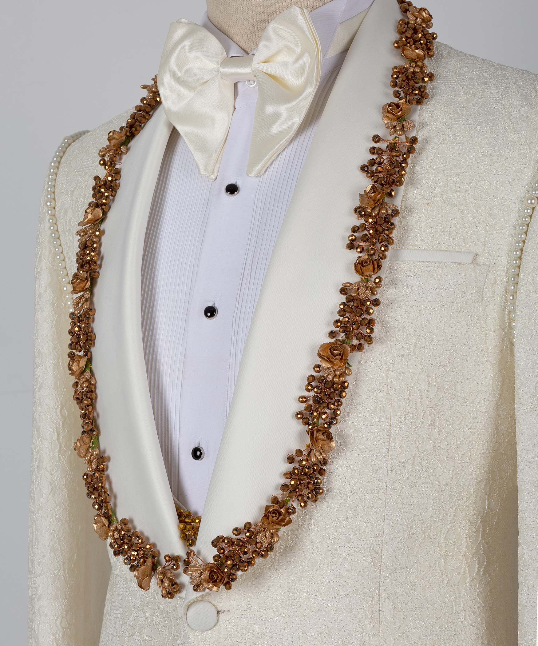 Close-up cream tuxedo jacket with gold beaded trim, white bow tie, and shirt placket
