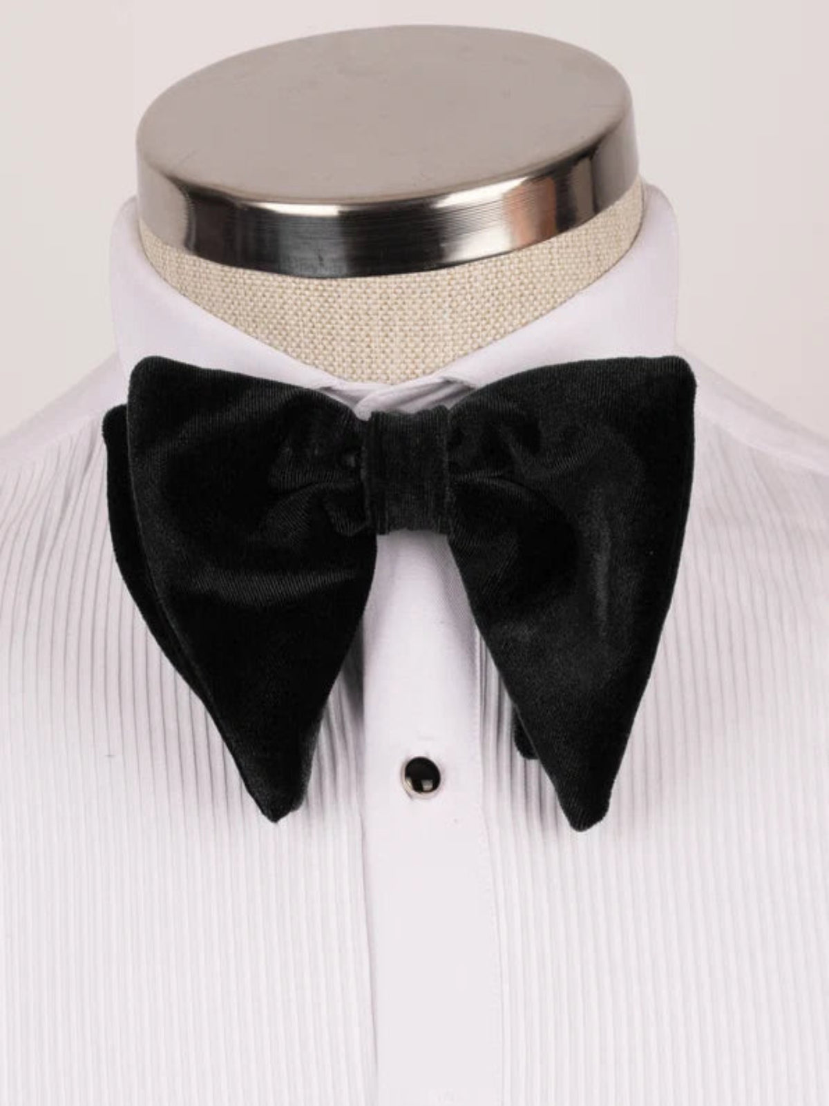 A black velvet bow tie displayed on a white pleated dress shirt mannequin with a silver neck cap.