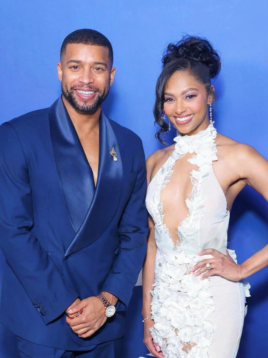 A man in a navy blue satin lapel suit and a woman in a white ruffled halter gown posing together against a solid blue backdrop.
