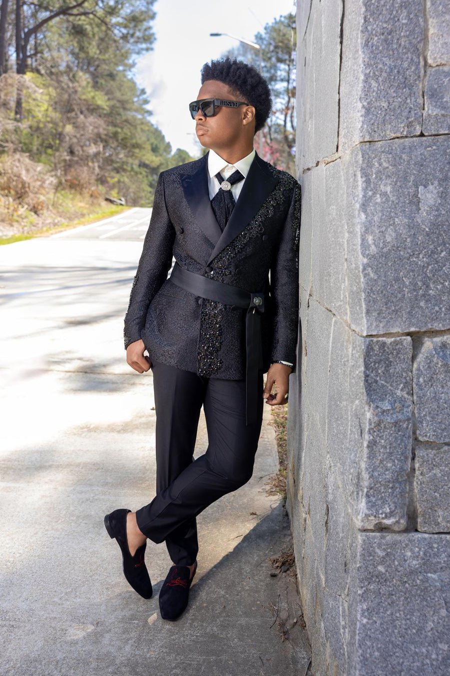 A man wearing a black double-breasted suit with textured lapels, a black waist belt, a white shirt, and a black tie leans against a gray stone wall on a paved road.