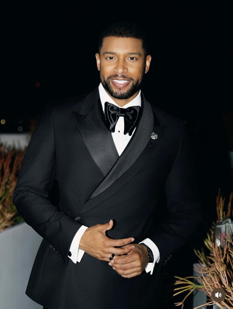 A man wearing a black tuxedo with a black satin bow tie and a white dress shirt, standing against a dark background with decorative plants at the sides.