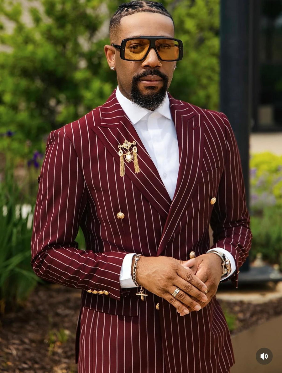 A man wearing a burgundy double-breasted suit with thin gold pinstripes, a white dress shirt, and oversized black sunglasses with amber lenses, standing outdoors in front of a blurred garden background.