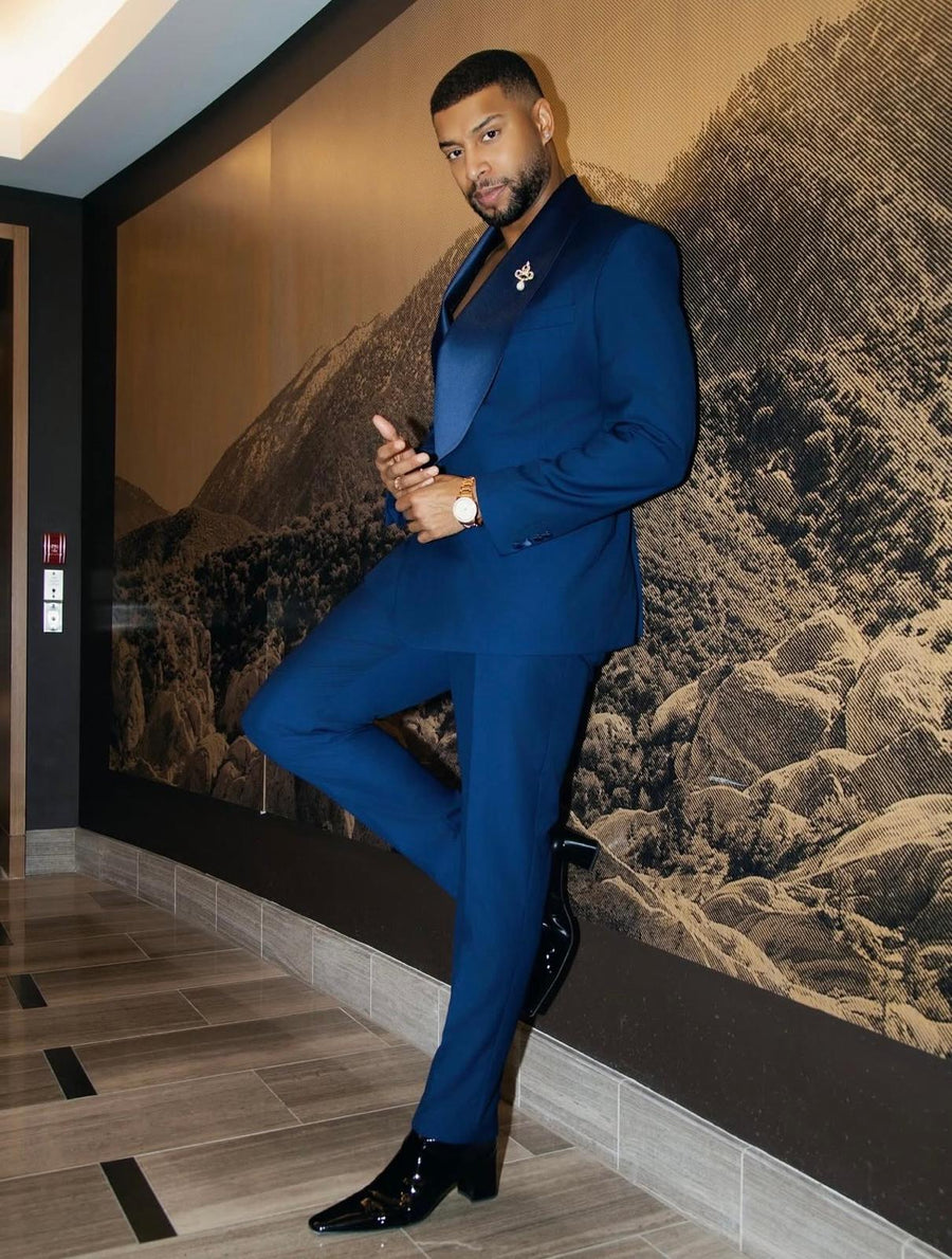 A man wearing a sharp royal blue two-piece suit with a matching blue shirt, black patent leather shoes, and a blue pocket square, posing in front of a large brown and black mountain mural.