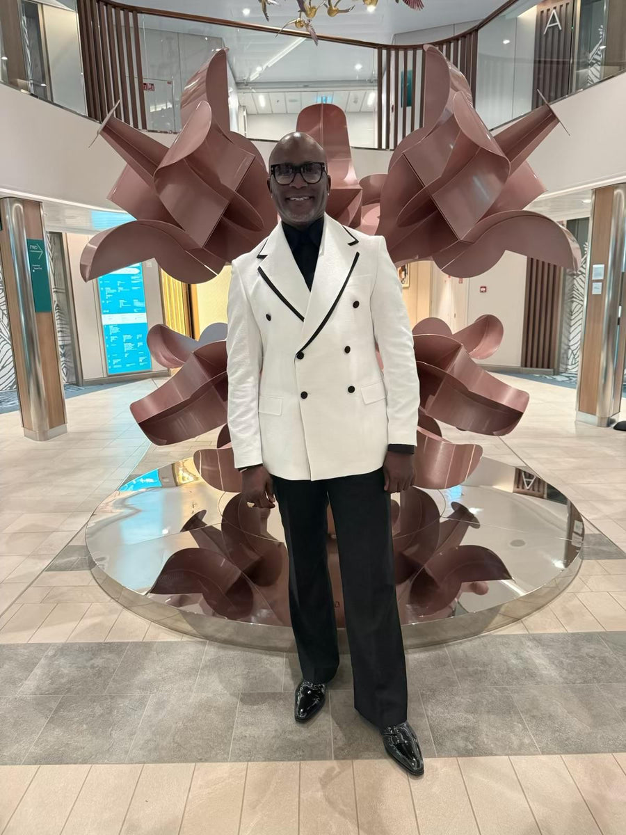 A man wearing a white double-breasted blazer with black trim, black trousers, and black shoes stands in front of a large bronze abstract sculpture in a modern indoor mall setting.