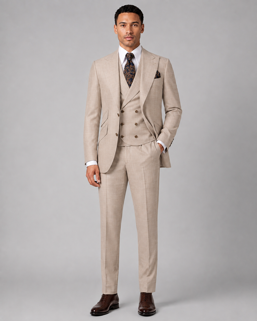 A man modeling a light beige three-piece suit with a double-breasted jacket, matching vest and trousers, a white dress shirt, a patterned tie, and brown dress shoes 