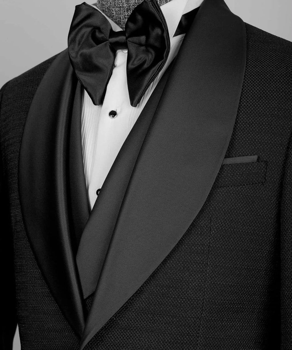 A close-up of a formal tuxedo featuring a black textured jacket with wide black satin shawl lapels, a white pleated dress shirt, a black satin bow tie, and a matching black vest.