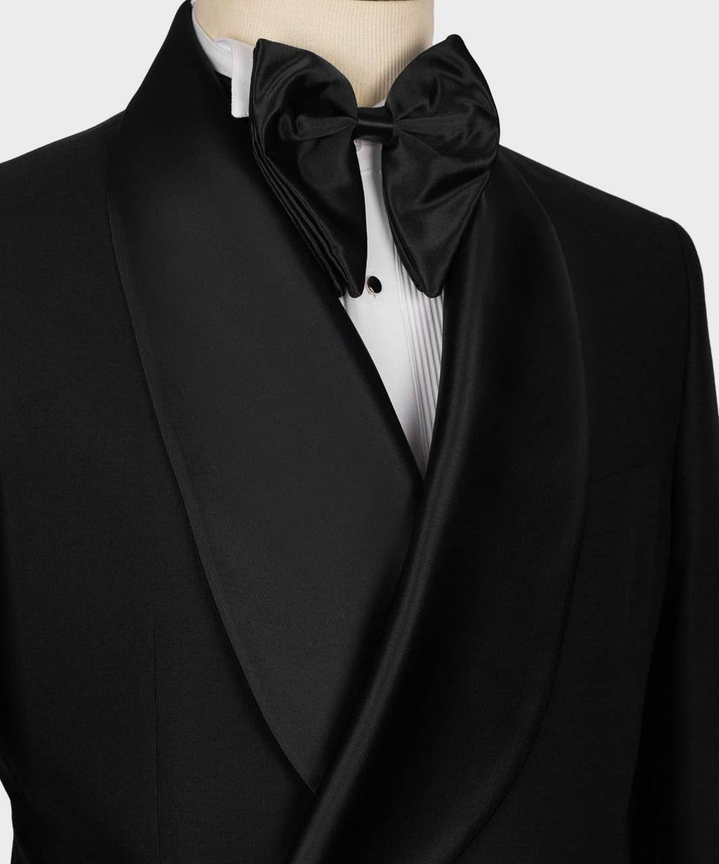 A close-up of a black tuxedo jacket with satin shawl lapels, worn with a white pleated dress shirt and a black satin bow tie on a mannequin torso.