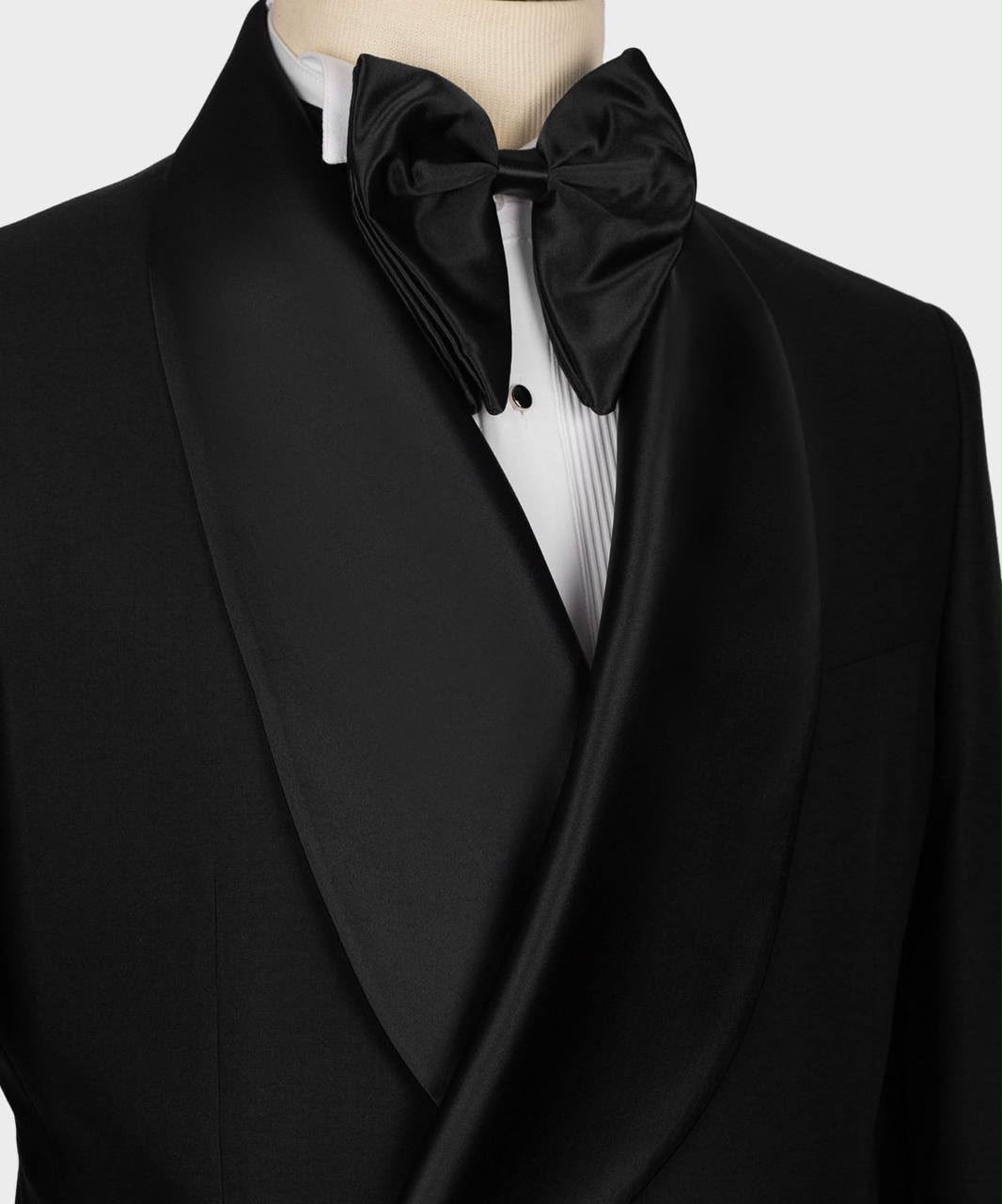 A close-up of a black tuxedo jacket with satin shawl lapels, worn with a white pleated dress shirt and a black satin bow tie on a mannequin torso.