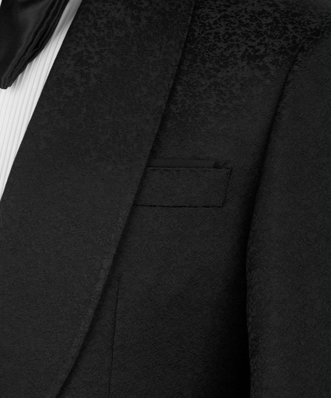 Close-up of a black textured tuxedo jacket lapel and chest pocket, paired with a white pleated shirt and black bow tie.