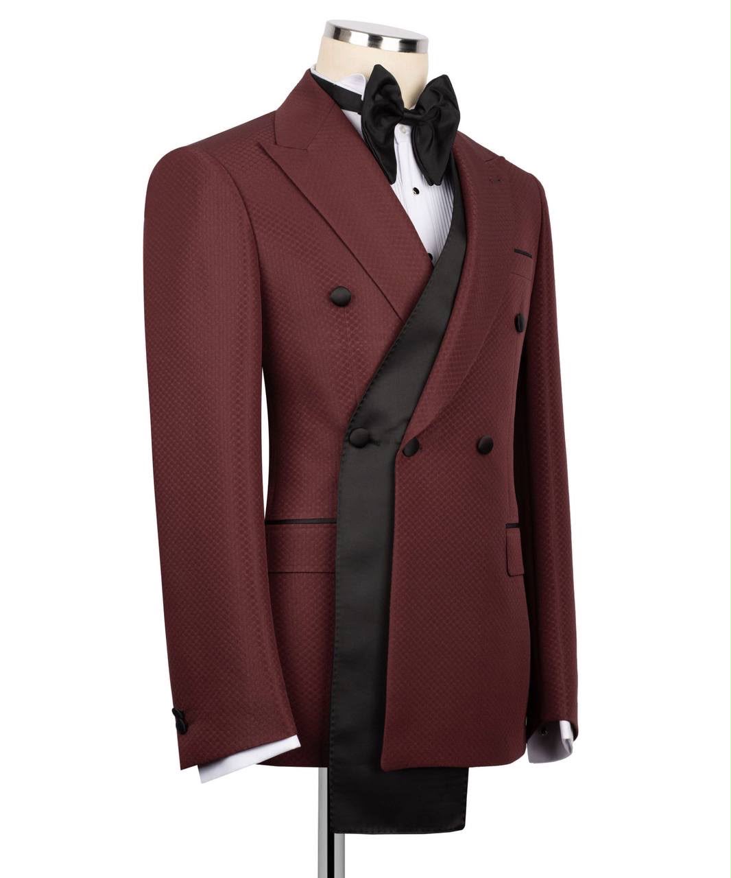 A men's double-breasted tuxedo jacket in burgundy with a subtle micro-pattern, styled on a mannequin with a black satin peak lapel, black bow tie, white dress shirt, and black vest 
