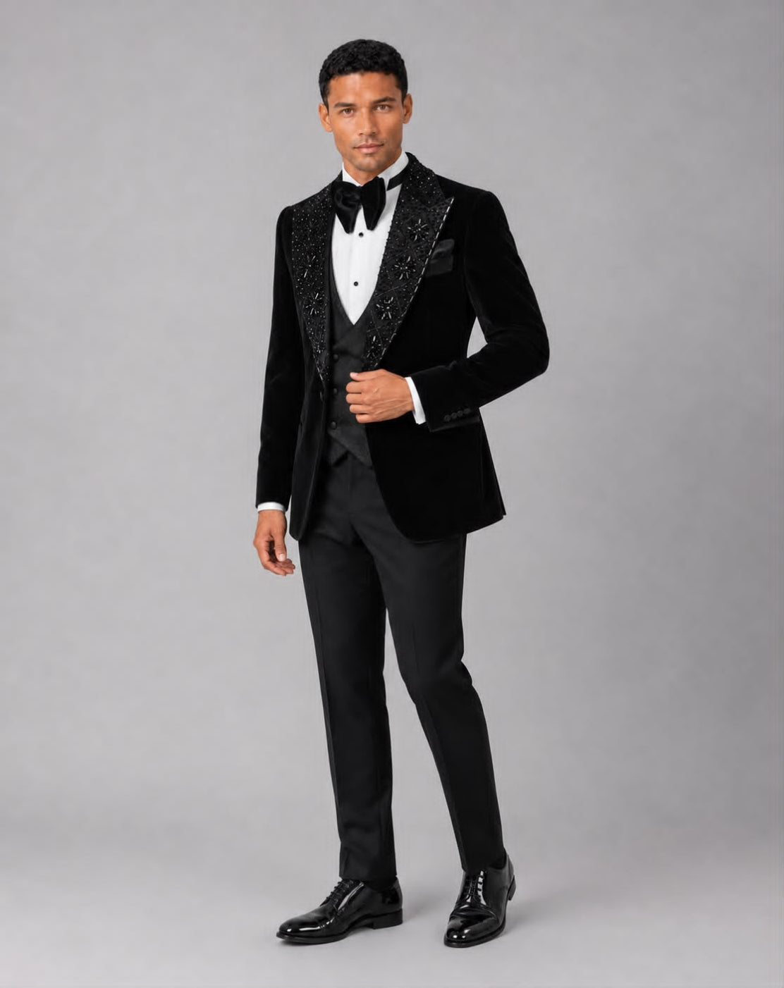 A man modeling a black velvet tuxedo jacket with sparkling beaded lapels, paired with a white dress shirt, black bow tie, black trousers, and black dress shoes, against a light gray studio background.