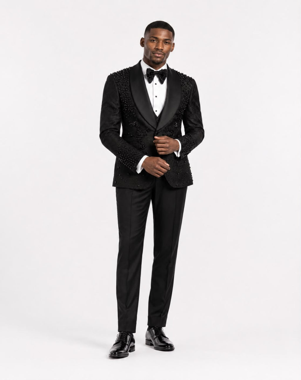 A man modeling a black tuxedo with ornate beaded lapels and shoulders, a white dress shirt, a black bow tie, and black trousers against a plain white studio background.