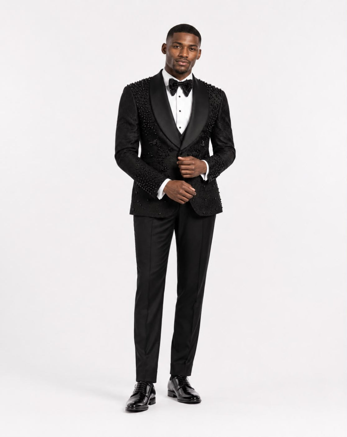 A man modeling a black tuxedo with ornate beaded lapels and shoulders, a white dress shirt, a black bow tie, and black trousers against a plain white studio background.