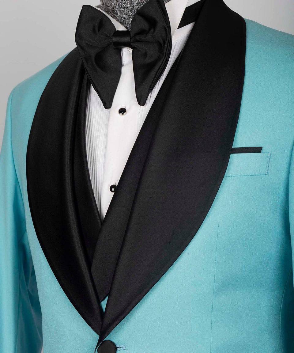 A close-up of a men's tuxedo jacket in bright turquoise with wide black satin shawl lapels, paired with a white pleated dress shirt, black bow tie, and black pocket square.
