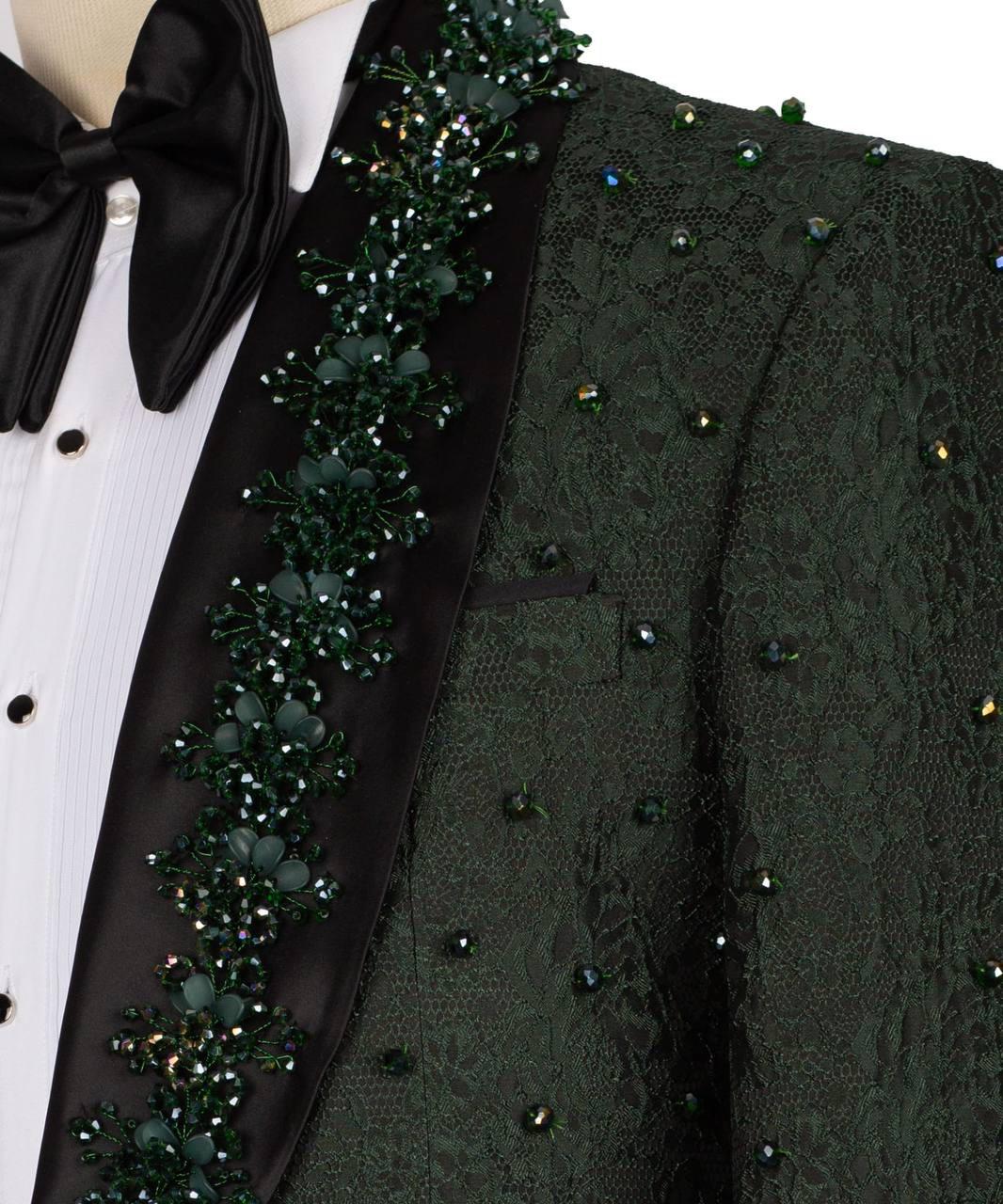 Close-up of a dark green lace tuxedo jacket with a black satin lapel and a decorative green floral beaded trim, worn over a white pleated shirt with a black bow tie.