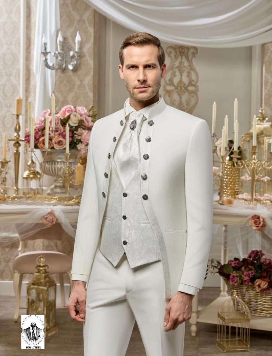 A man wearing a formal white three-piece suit with a high collar, double-breasted jacket, matching vest, and tie, standing in an ornate, elegant room decorated with gold candelabras and pink flowers.