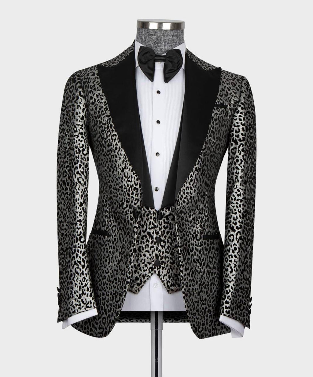 A men's tuxedo jacket with a black and gray leopard print pattern and black satin peak lapels, displayed on a mannequin with a white dress shirt, black bow tie, and white cufflinks.