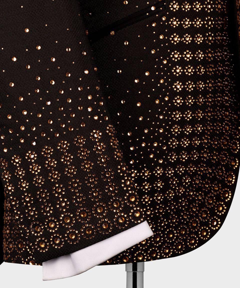 Close-up of a dark brown or black suit jacket heavily embellished with gold-toned studs and rhinestones, with a white shirt cuff visible at the hem.