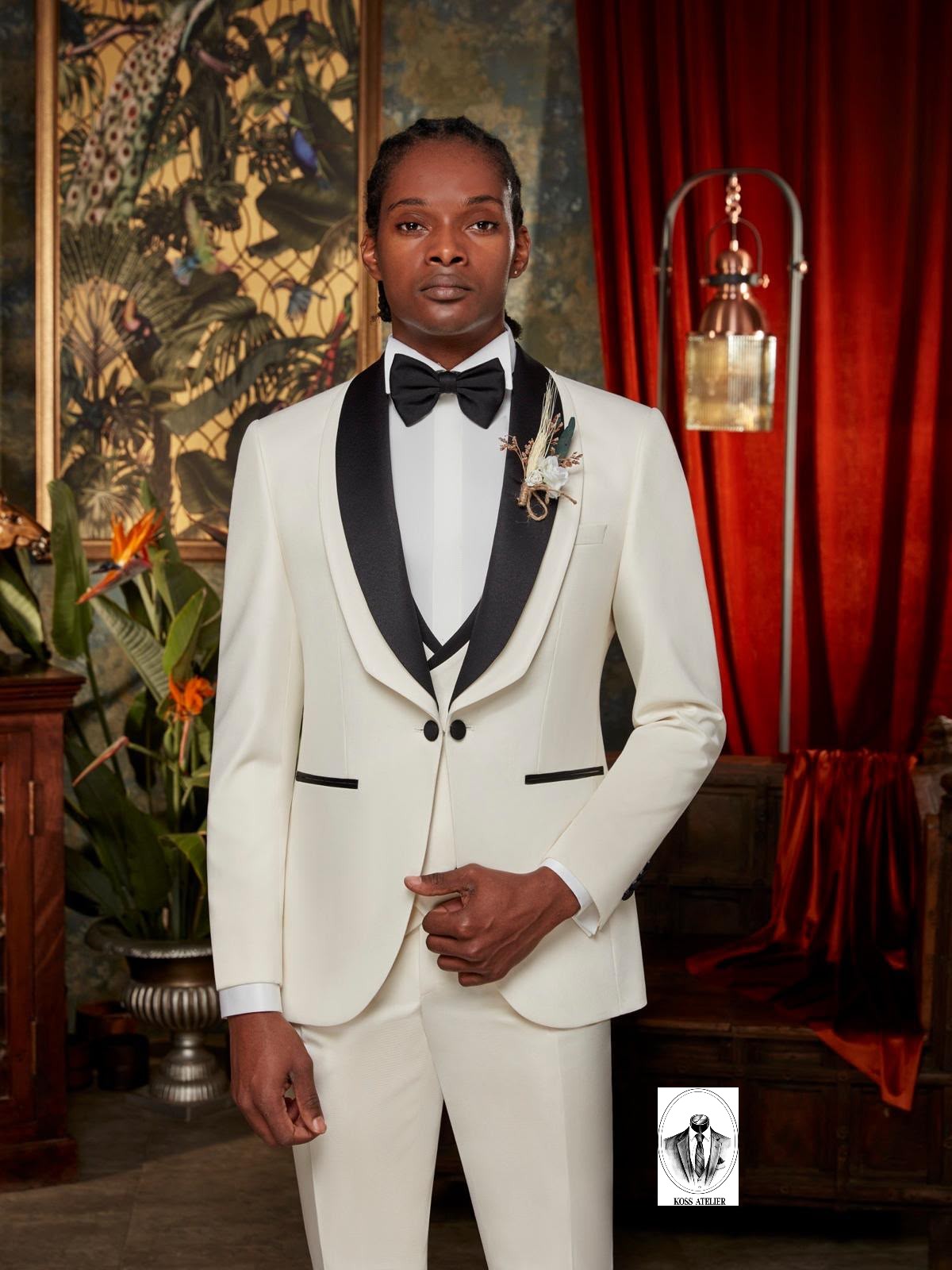 A man wearing a white tuxedo with black satin lapels, a black bow tie, and a floral boutonniere, standing in a decorated room with red curtains and a large tapestry.