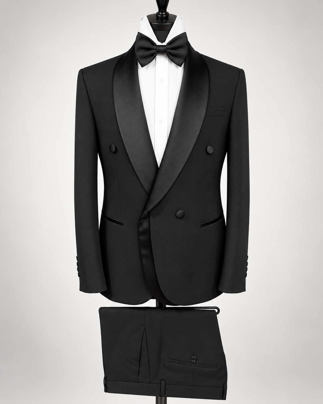 A black double-breasted tuxedo jacket with satin shawl lapels, displayed on a mannequin with a white dress shirt and black bow tie. Matching black trousers are shown below on a stand against a light gray studio background.