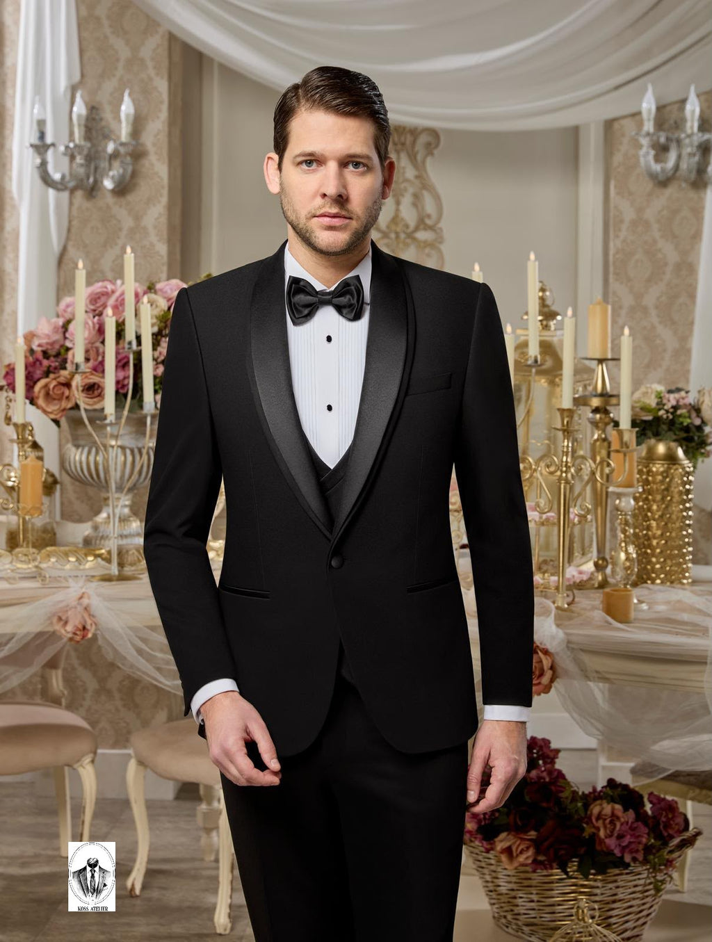 A man wearing a classic black tuxedo with satin peak lapels, a white dress shirt, and a black bow tie, standing in an elegant, decorated event hall with gold candelabras and floral arrangements.