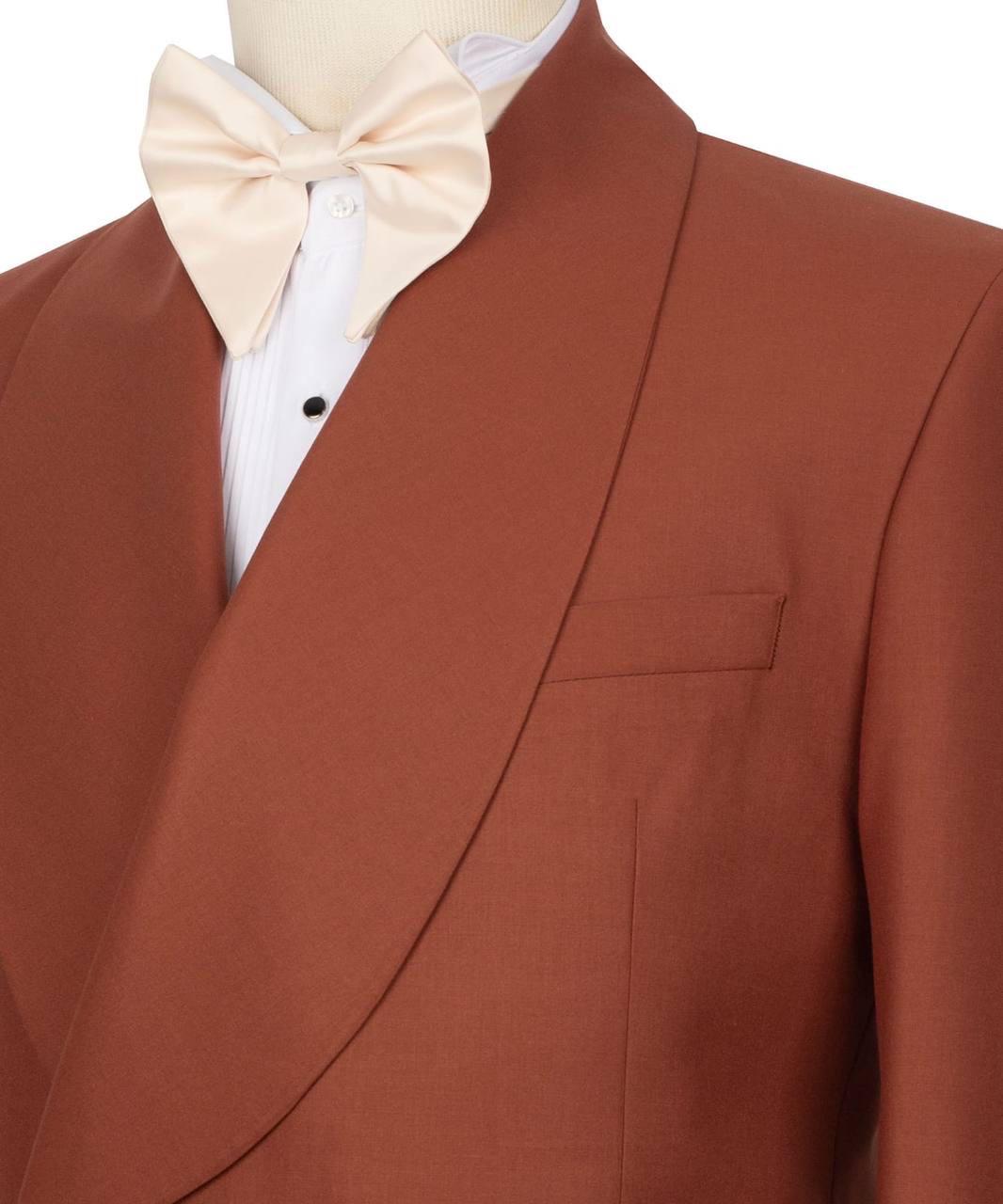 A close-up of a men's formalwear outfit on a mannequin, featuring a rust-brown shawl-collar tuxedo jacket over a white pleated tuxedo shirt with a cream bow tie.