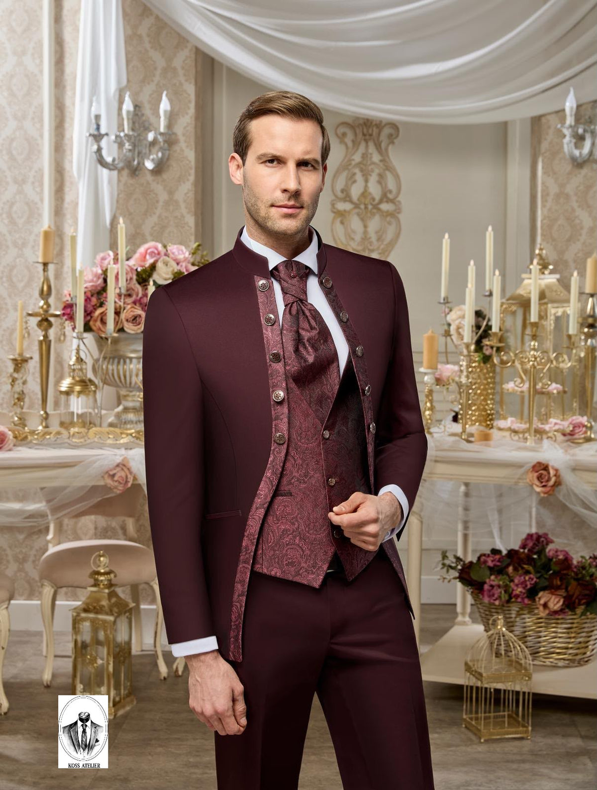 A man modeling a three-piece burgundy formal suit with a matching patterned vest, tie, and trousers, set against an elegant, vintage-style event venue with draped white fabric, gold tables, and floral arrangements.