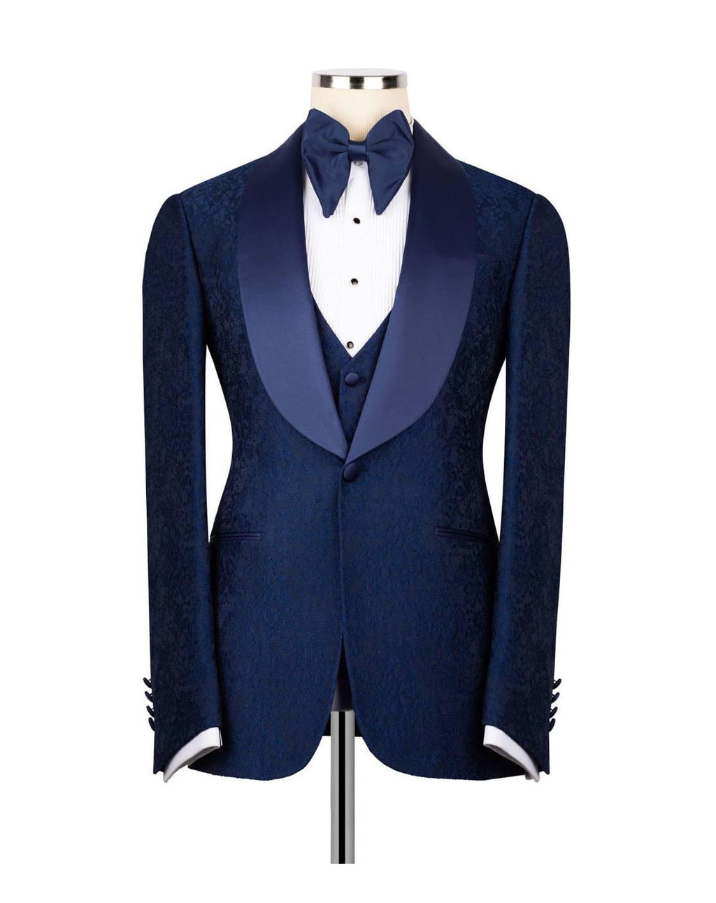 A men's formal tuxedo jacket displayed on a mannequin torso, featuring a textured navy blue body, satin peak lapels, a matching navy blue bow tie, white dress shirt, and a navy blue vest.