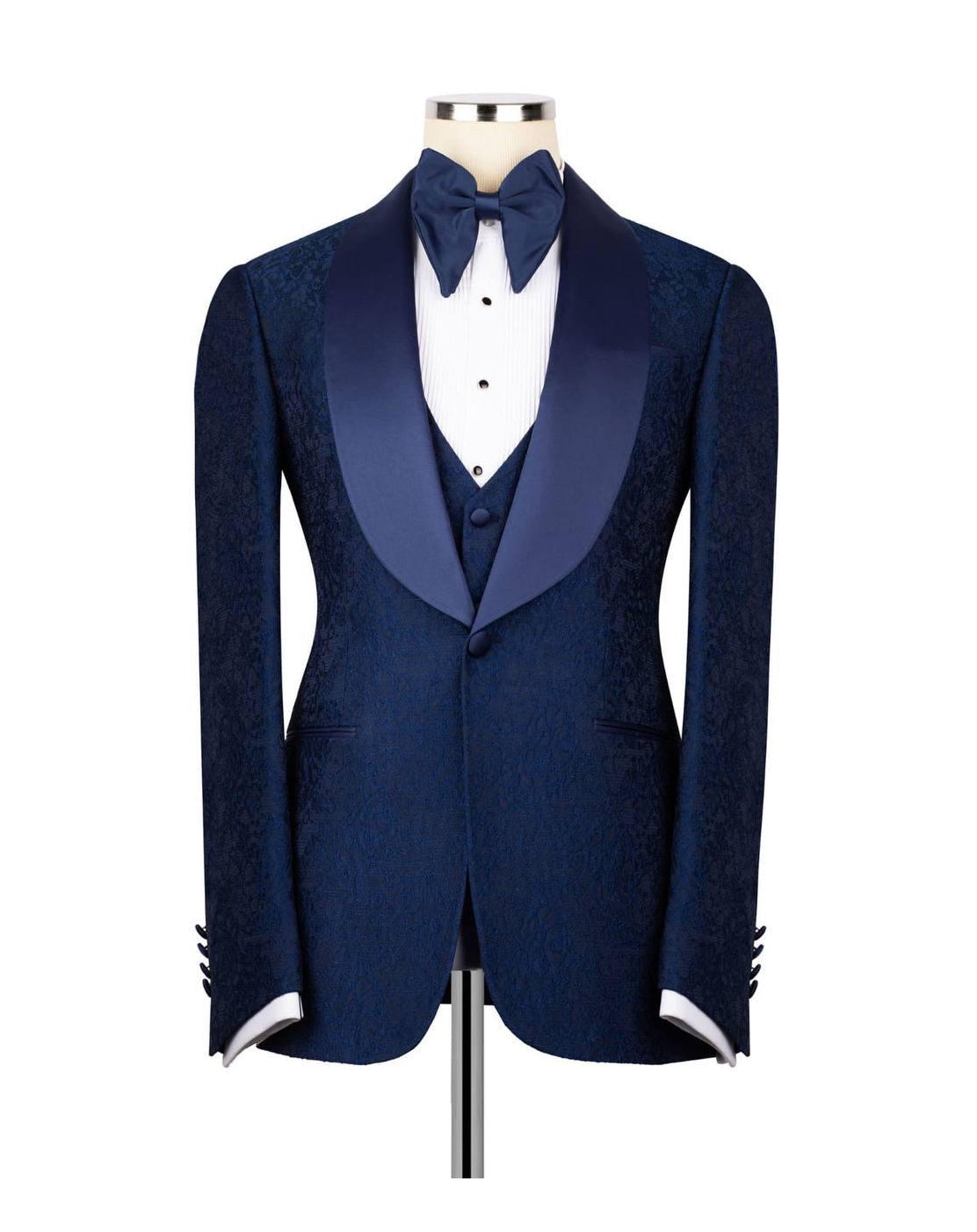 A men's formal tuxedo jacket displayed on a mannequin torso, featuring a textured navy blue body, satin peak lapels, a matching navy blue bow tie, white dress shirt, and a navy blue vest.