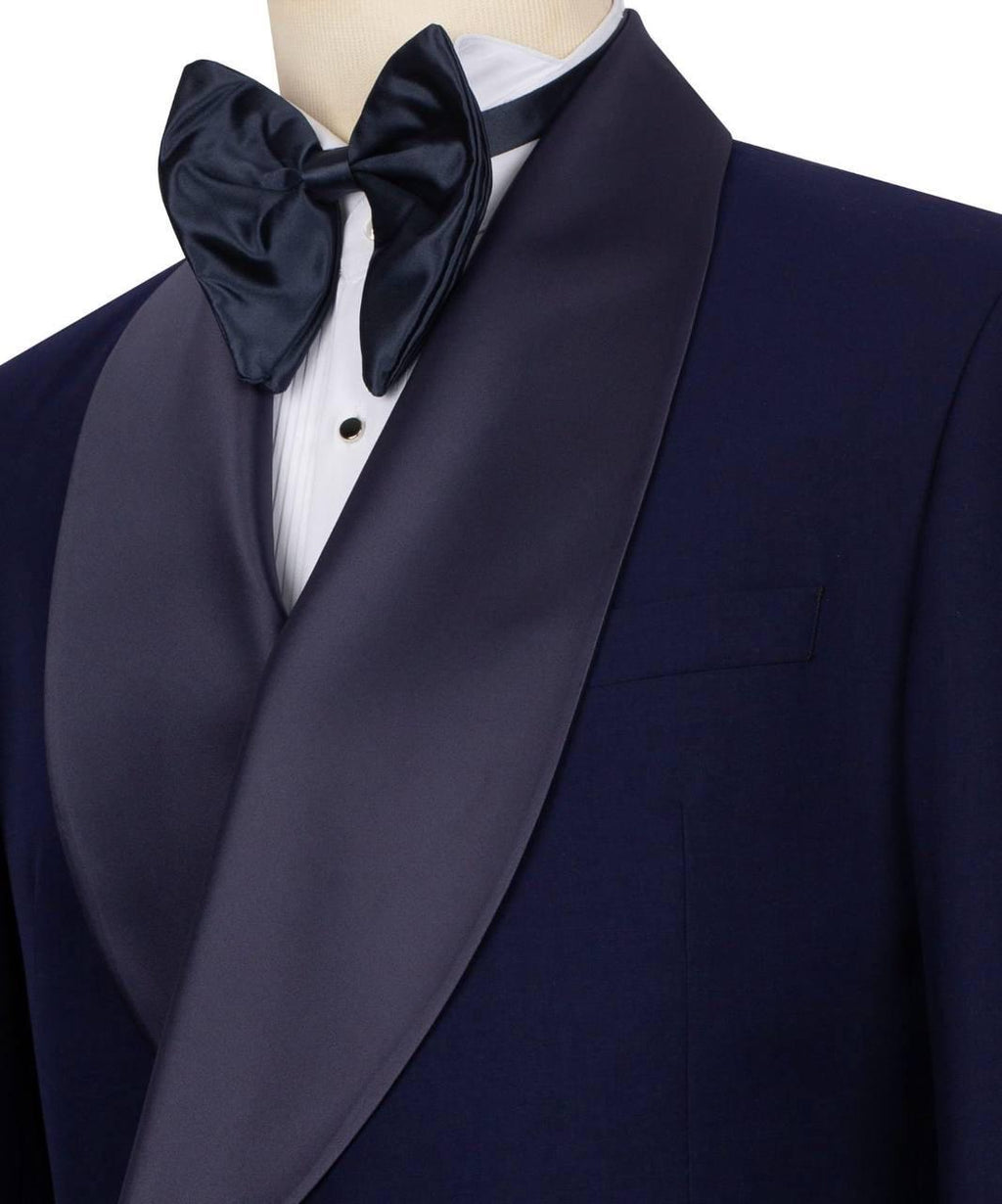 A close-up of a formal tuxedo jacket with dark navy satin shawl lapels, worn over a white dress shirt and paired with a matching navy satin bow tie on a mannequin.