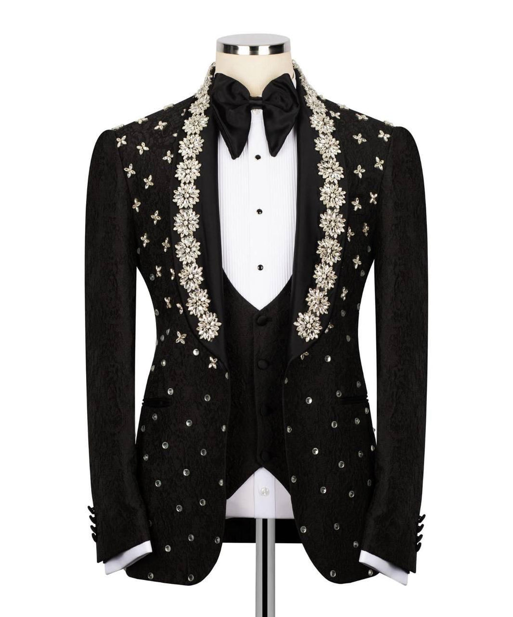 A black tuxedo jacket with ornate silver and white floral embroidery on the lapels and scattered silver embellishments on the front, displayed on a mannequin with a white tuxedo shirt, black vest, black bow tie, and black trousers against a white background.
