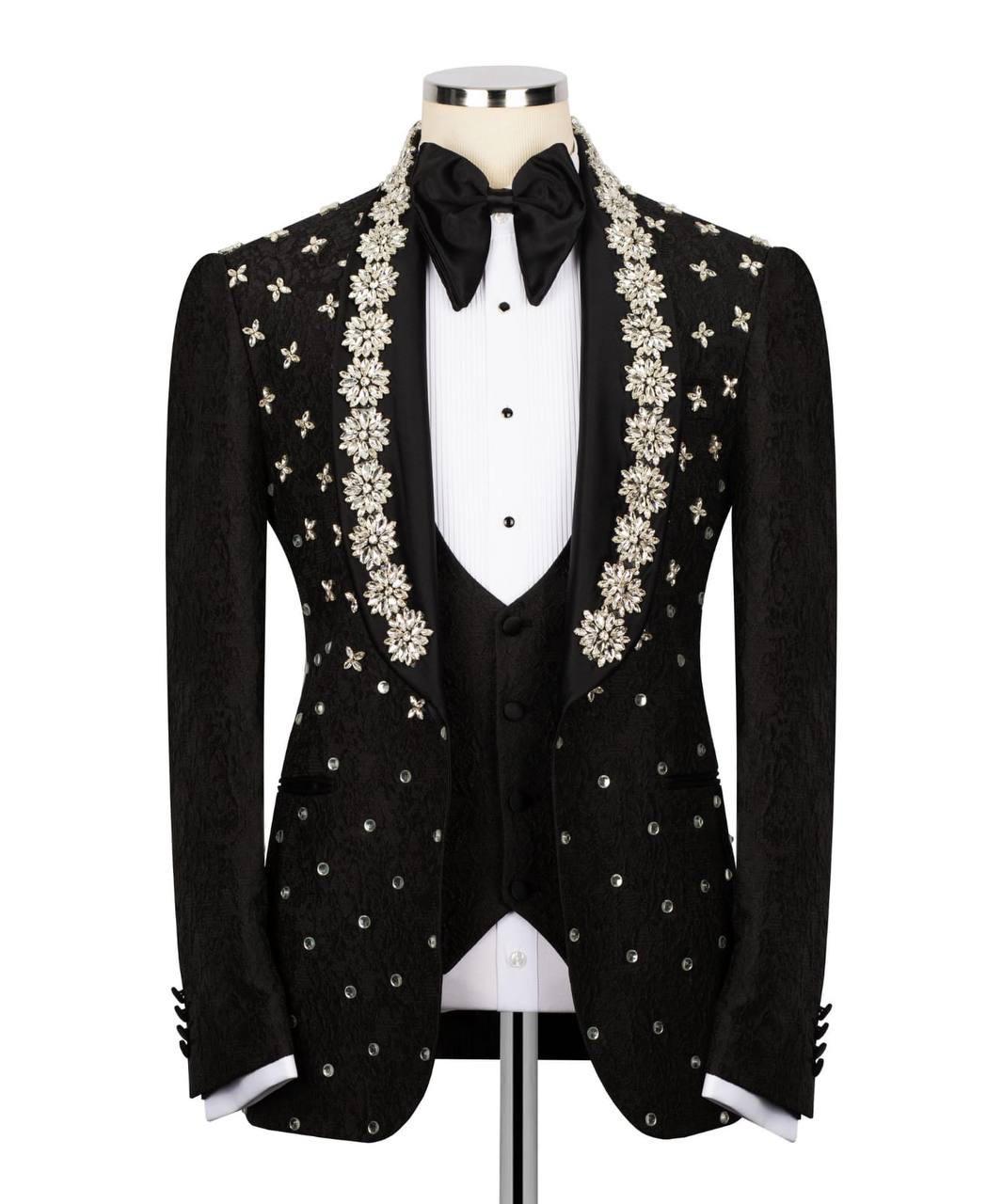 A black tuxedo jacket with ornate silver and white floral embroidery on the lapels and scattered silver embellishments on the front, displayed on a mannequin with a white tuxedo shirt, black vest, black bow tie, and black trousers against a white background.