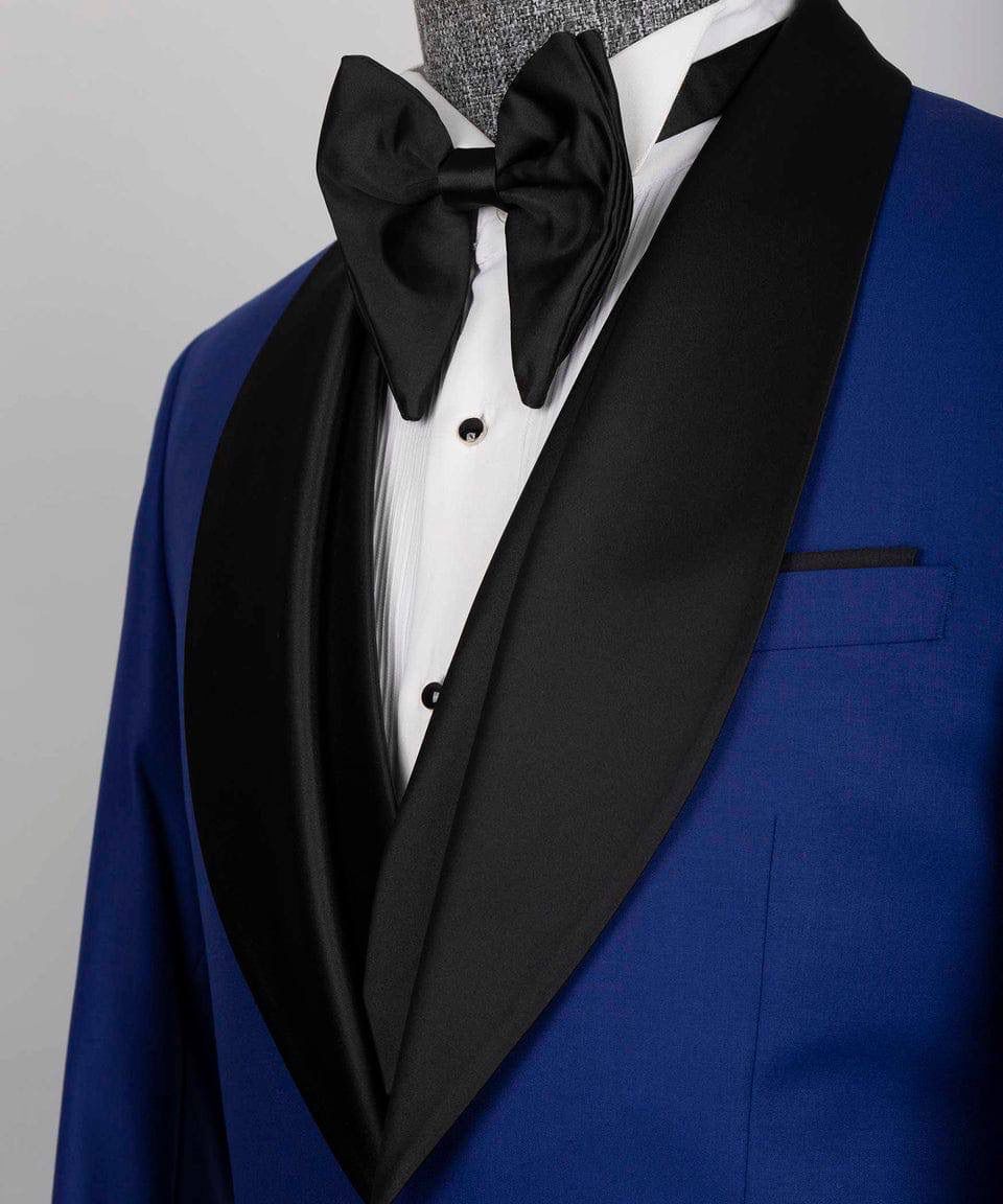 A close-up of a men's tuxedo jacket in royal blue with black satin shawl lapels, styled with a white dress shirt, black bow tie, and a gray textured mannequin neck.