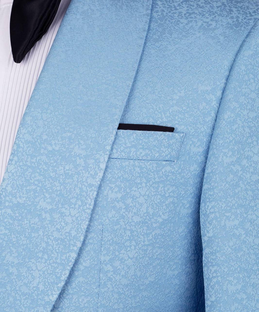 A close-up of a light blue textured suit jacket with a subtle damask pattern, featuring a black pocket square and a white pleated shirt underneath.