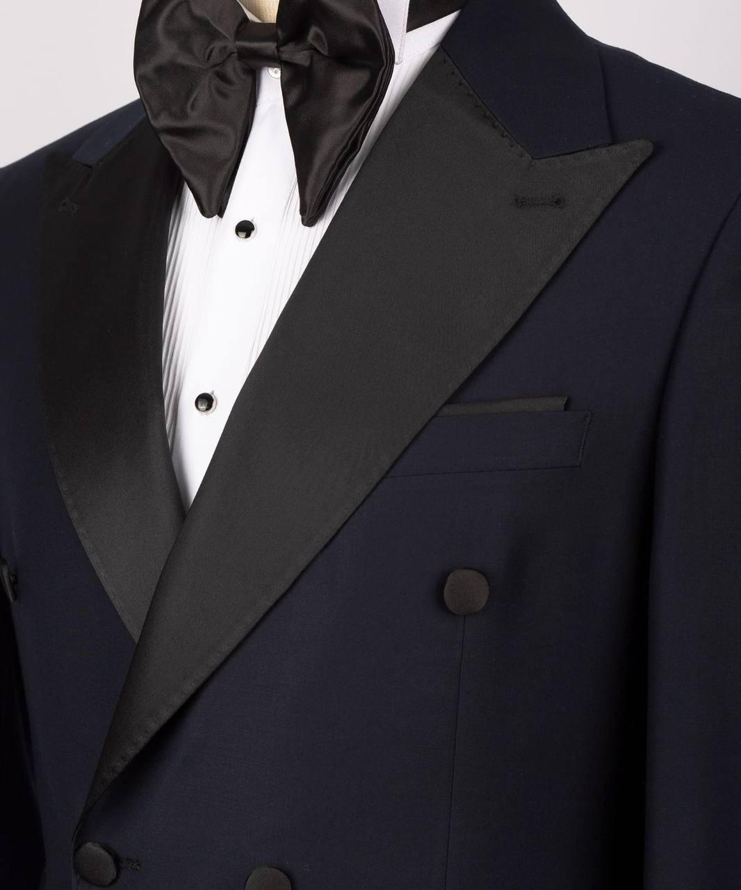 Close-up of a formal tuxedo jacket in dark navy with black satin lapels, worn with a white pleated shirt and black bow tie.