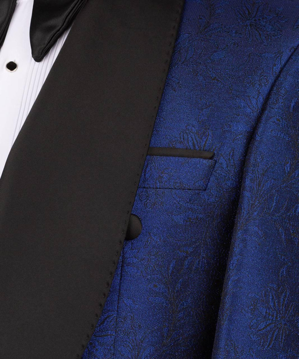 Close-up of a formal tuxedo jacket featuring a rich royal blue brocade patterned vest and contrasting black satin lapels, worn over a white pleated shirt and black bow tie.