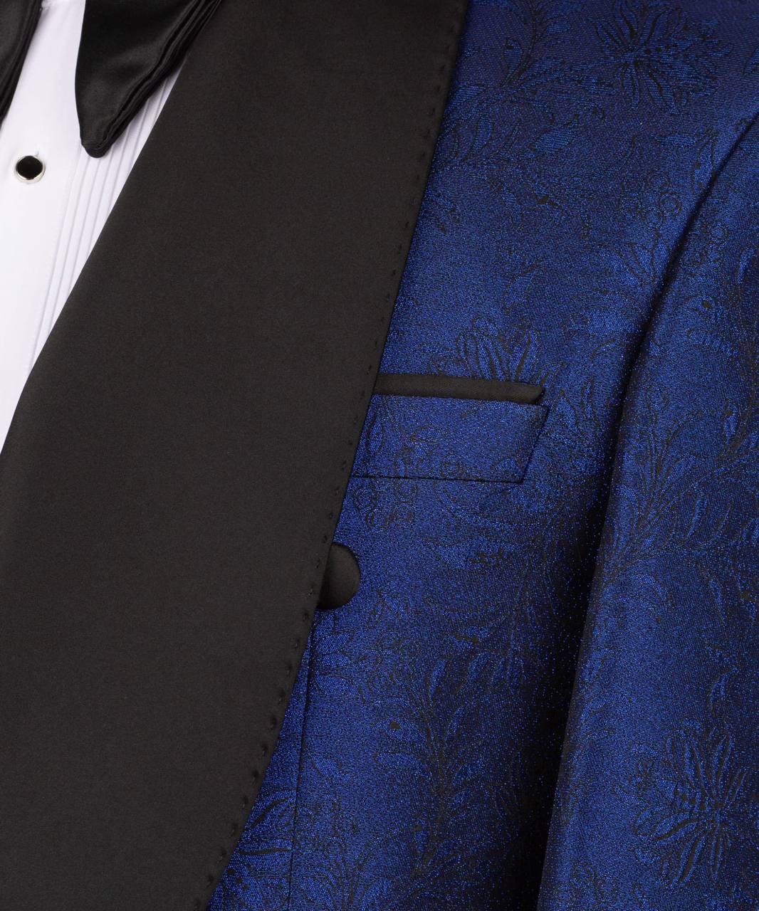 Close-up of a formal tuxedo jacket featuring a rich royal blue brocade patterned vest and contrasting black satin lapels, worn over a white pleated shirt and black bow tie.