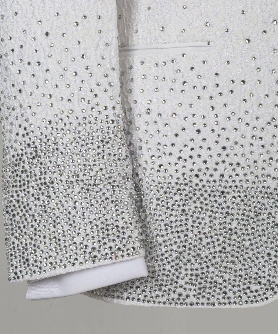 Close-up of a white textured jacket or blazer heavily embellished with silver rhinestones, showing the sleeve cuff and front panel against a neutral gray background.