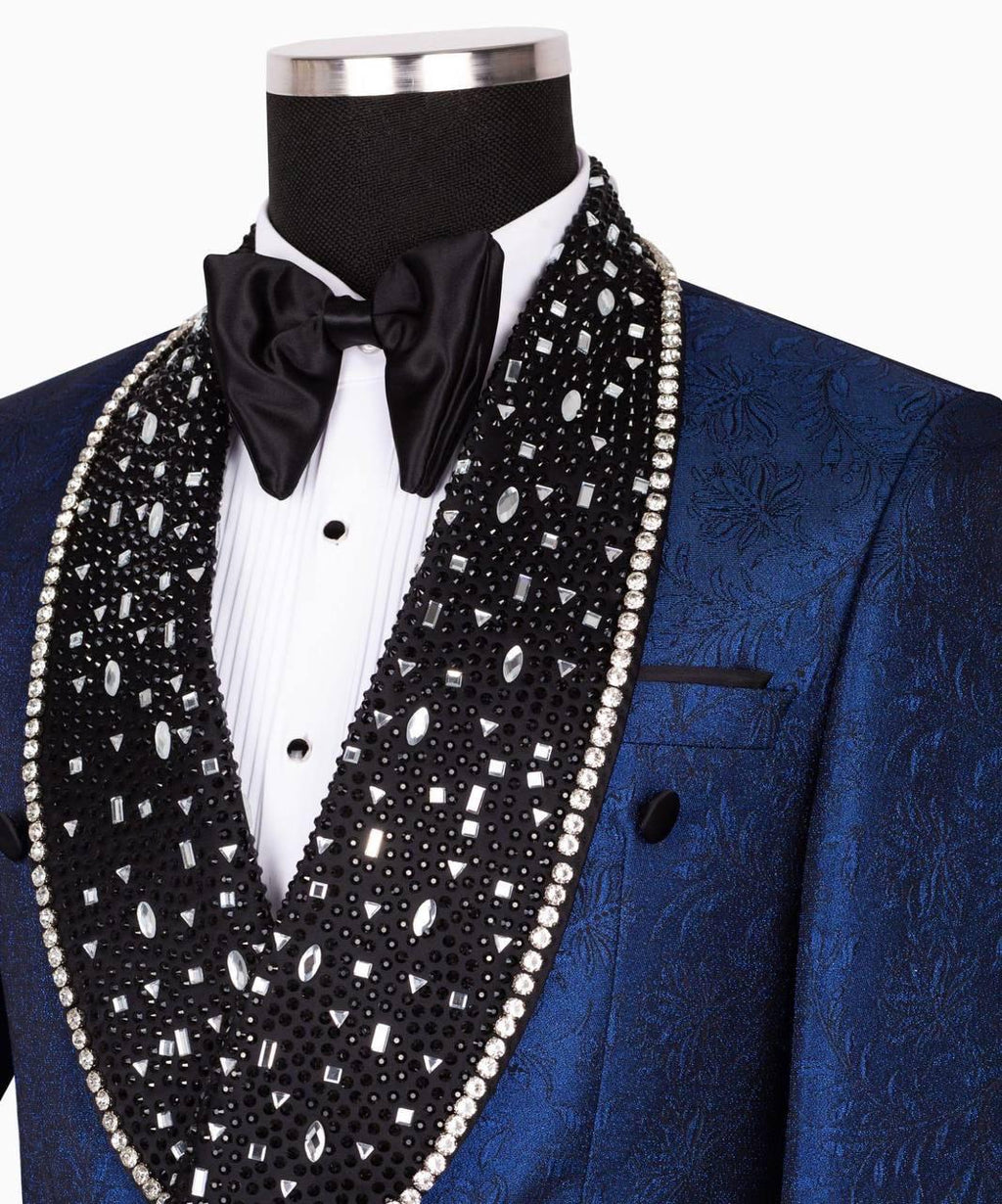 A close-up of a blue textured tuxedo jacket displayed on a mannequin torso, featuring a black satin bow tie and a large black shawl vest embellished with silver rhinestones and white faux pearls.