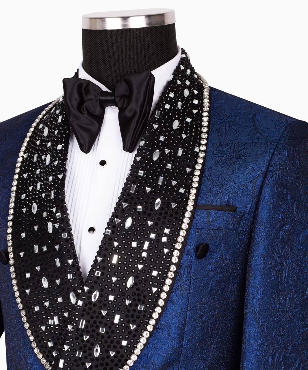 A close-up of a blue textured tuxedo jacket displayed on a mannequin torso, featuring a black satin bow tie and a large black shawl vest embellished with silver rhinestones and white faux pearls.