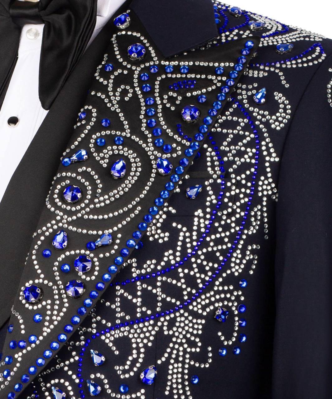 Close-up of a black tuxedo jacket lapel embellished with intricate silver rhinestone swirls and royal blue crystal accents, worn over a black tuxedo shirt and bow tie.