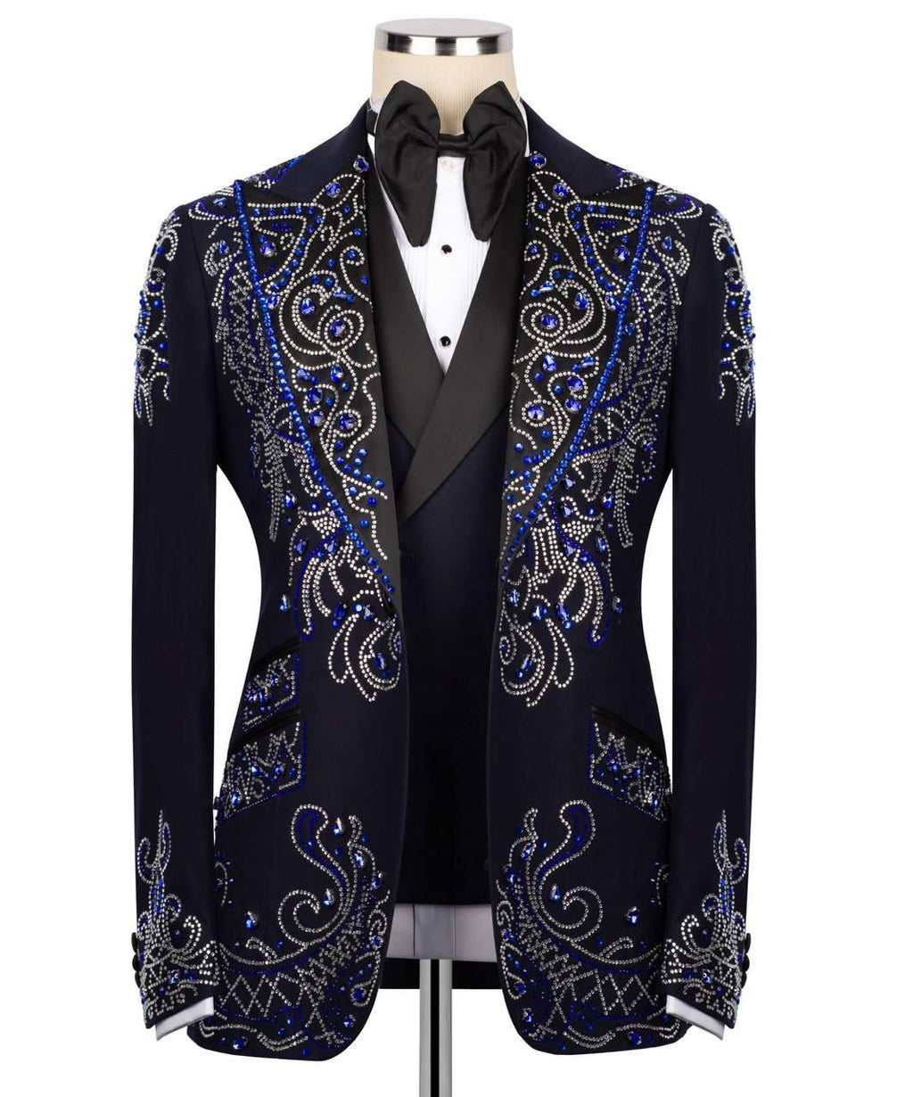 A men's formal blazer jacket displayed on a mannequin torso, featuring ornate silver and blue rhinestone paisley embroidery over a dark navy base. The jacket is styled with a black bow tie, white dress shirt, and black vest against a plain white background.