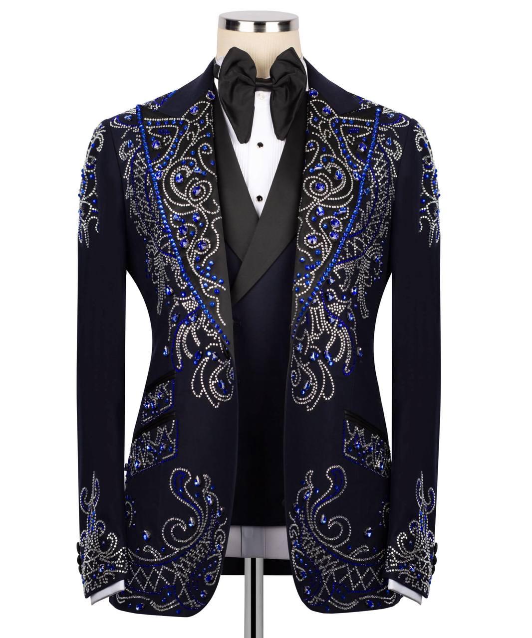 A men's formal blazer jacket displayed on a mannequin torso, featuring ornate silver and blue rhinestone paisley embroidery over a dark navy base. The jacket is styled with a black bow tie, white dress shirt, and black vest against a plain white background.