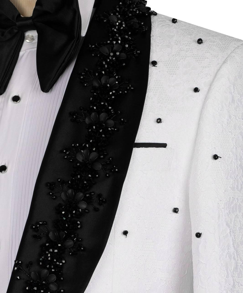 A close-up of a formal tuxedo jacket featuring a white textured lace body, a black satin peak lapel heavily embellished with black floral embroidery and beadwork, a black bow tie, and a white pleated dress shirt.