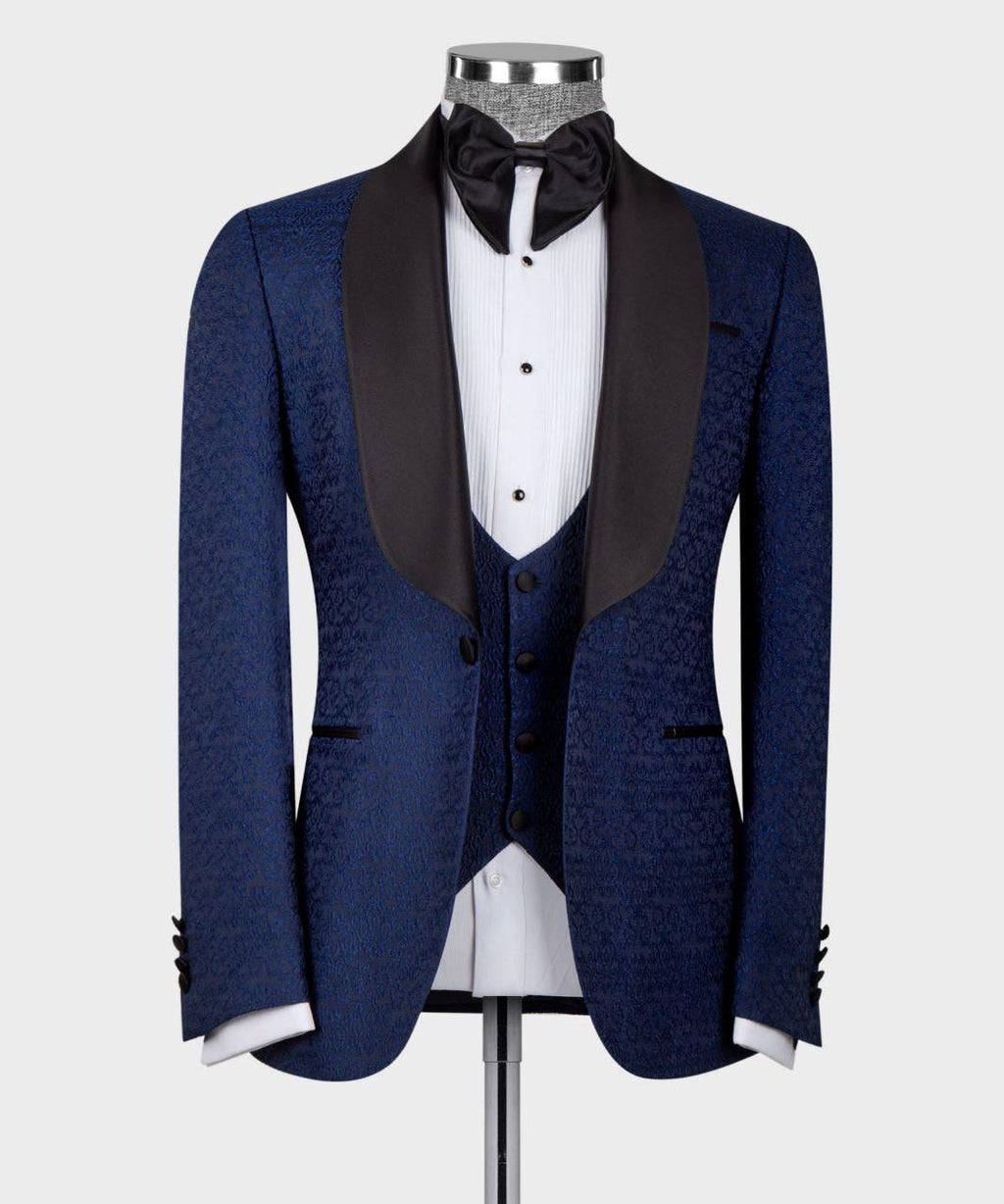 A men's tuxedo jacket displayed on a mannequin, featuring a textured navy blue patterned fabric with contrasting black satin shawl lapels, styled with a white pinstripe shirt, black bow tie, and matching navy vest.