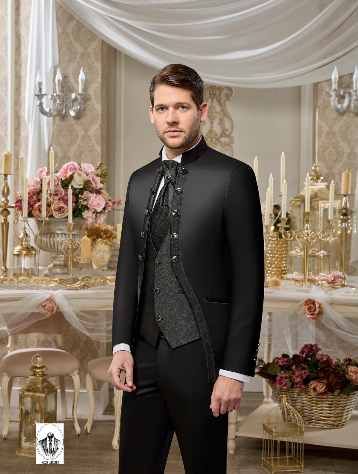 A man wearing a formal black suit with a high collar, matching vest, and patterned tie, standing in an elegant, vintage-style room decorated with draped white fabric, gold candle holders, and floral arrangements.