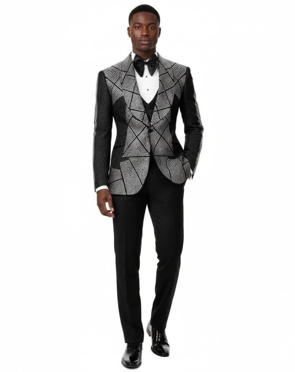 A man modeling a black and white geometric patterned tuxedo jacket with black satin peak lapels, paired with black trousers, a white shirt, and a black bow tie against a white background.