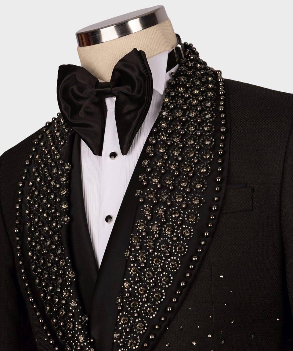 A close-up of a black tuxedo jacket with ornate silver and black beaded shawl lapels, styled with a white pinstripe shirt, black satin bow tie, and black vest on a mannequin torso.