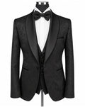 A men's formal tuxedo jacket with black satin shawl collar, black paisley brocade fabric body, paired with a matching black vest, black bow tie, and white dress shirt on a mannequin torso.