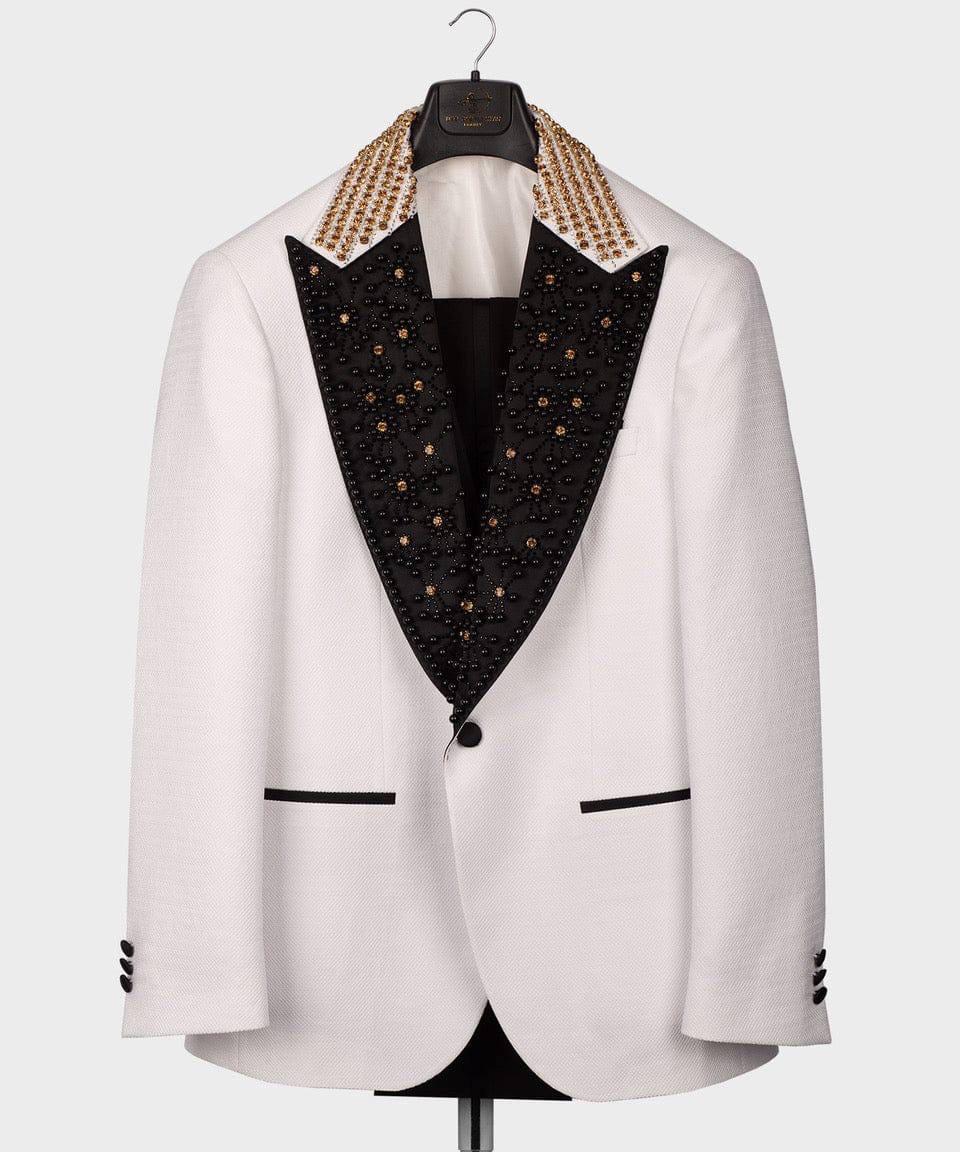 A white blazer jacket with a black lapel heavily embellished with gold studs and a matching gold beaded collar, displayed on a hanger against a light gray background.